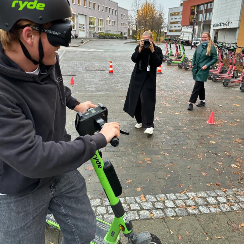 En mann med promillebriller og hjelp prøver å navigere seg gjennom en løype med kjegler mens han kjører elsparkesykkel. Vi ser astfalt i bakgrunnen og to personer som ser på. Det står parkert flere elsparkesykler lenger bak.