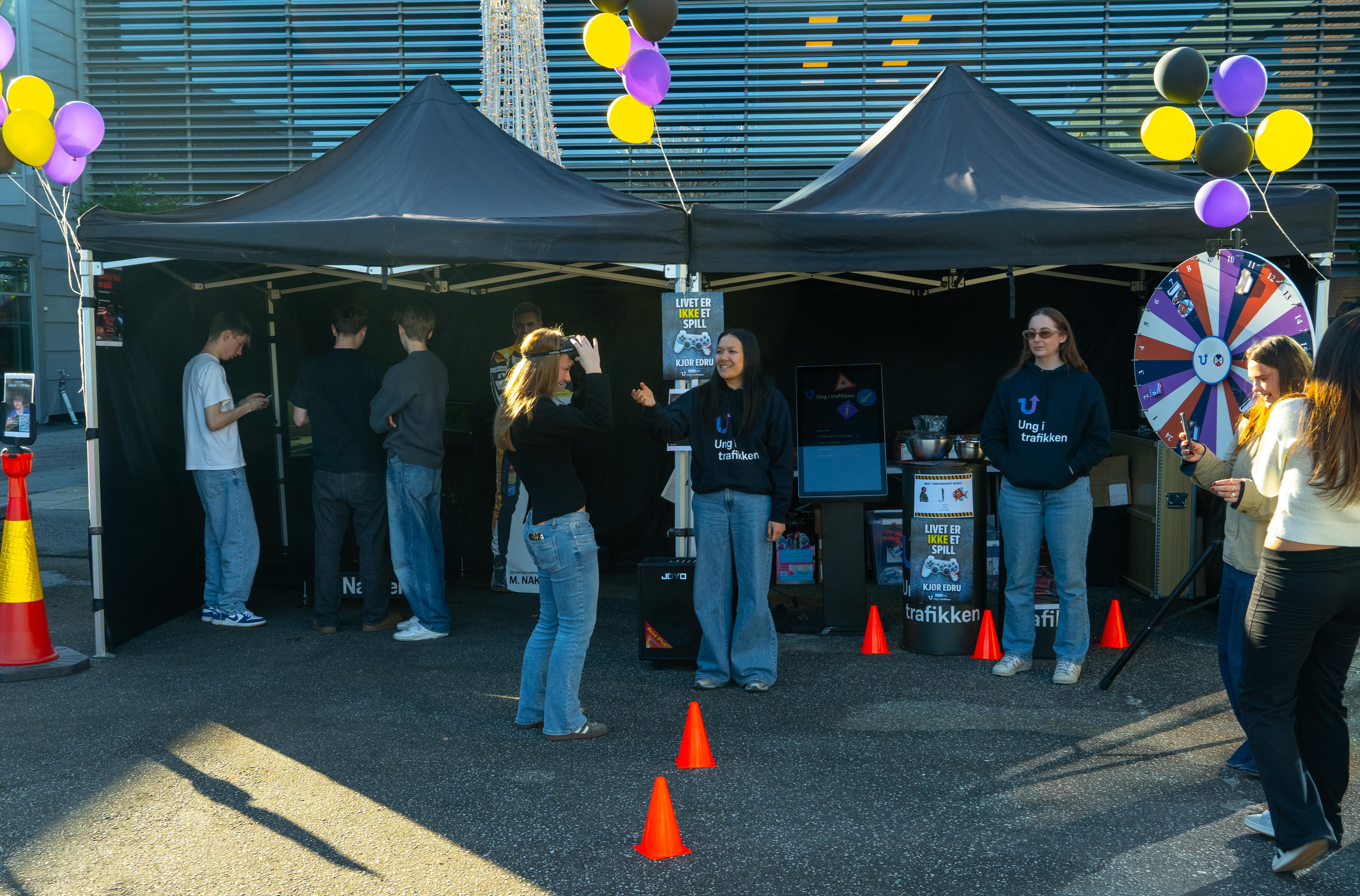 Utstilling bestående av to svarte telt med fargede ballonger som dekorasjon. Flere ungdommer som står foran og tester ut promillebriller mens de går gjennom en bane med kjegler. Lykkehjulet står klart for å brukes.