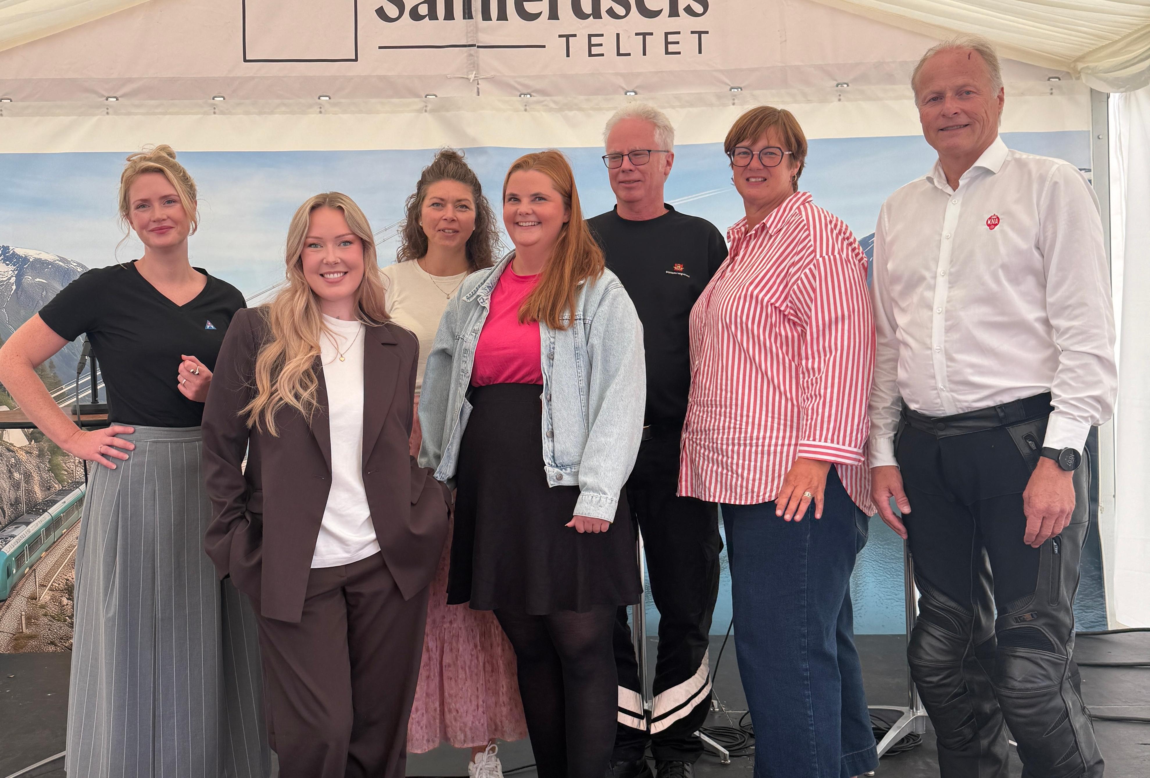 Bildet viser 6 personerstå ved siden av hverandre i et hvitt telt på en svart scene. De smiler til kamera.