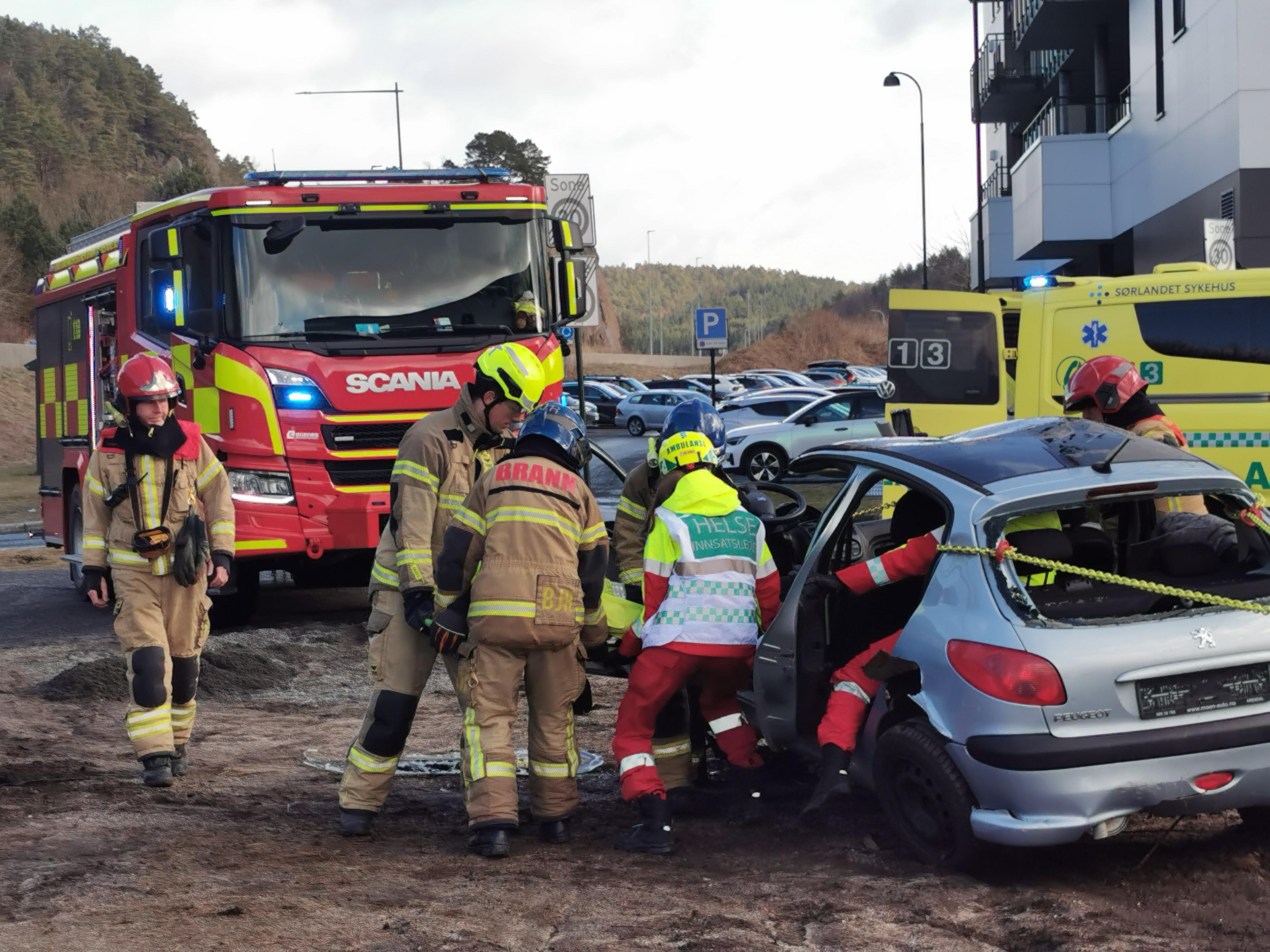 Bildet viser rød brannbil og gul ambulanse i bakgrunn. I front ser vi 3 brannbetjente og to ambulansearbeidere ta ut pasienten fra bilen.
