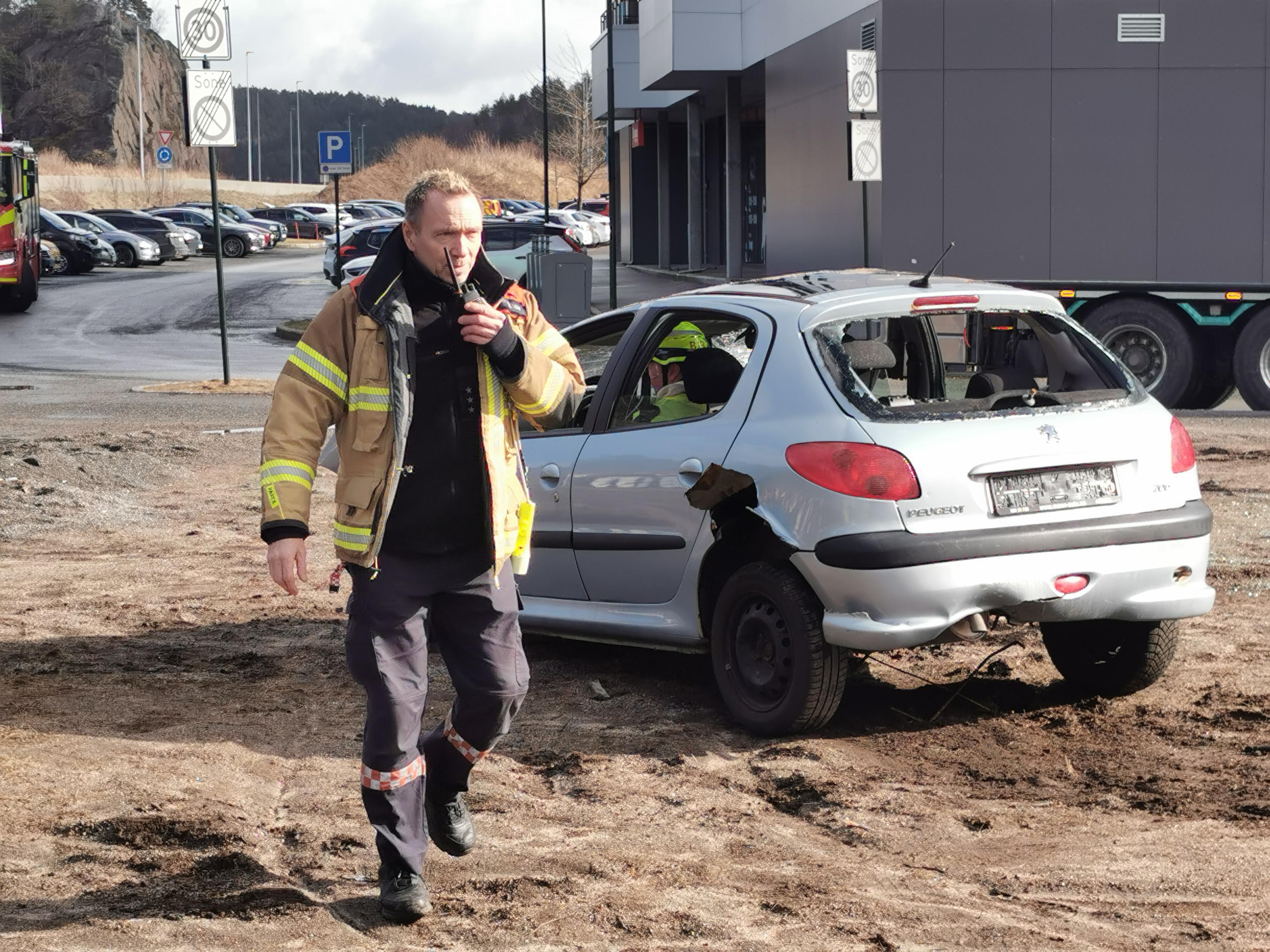 En brannmann står foran bilen som droppet og prater i walk-e-talkie. Bilen er sølvfarget og knust i fronten.