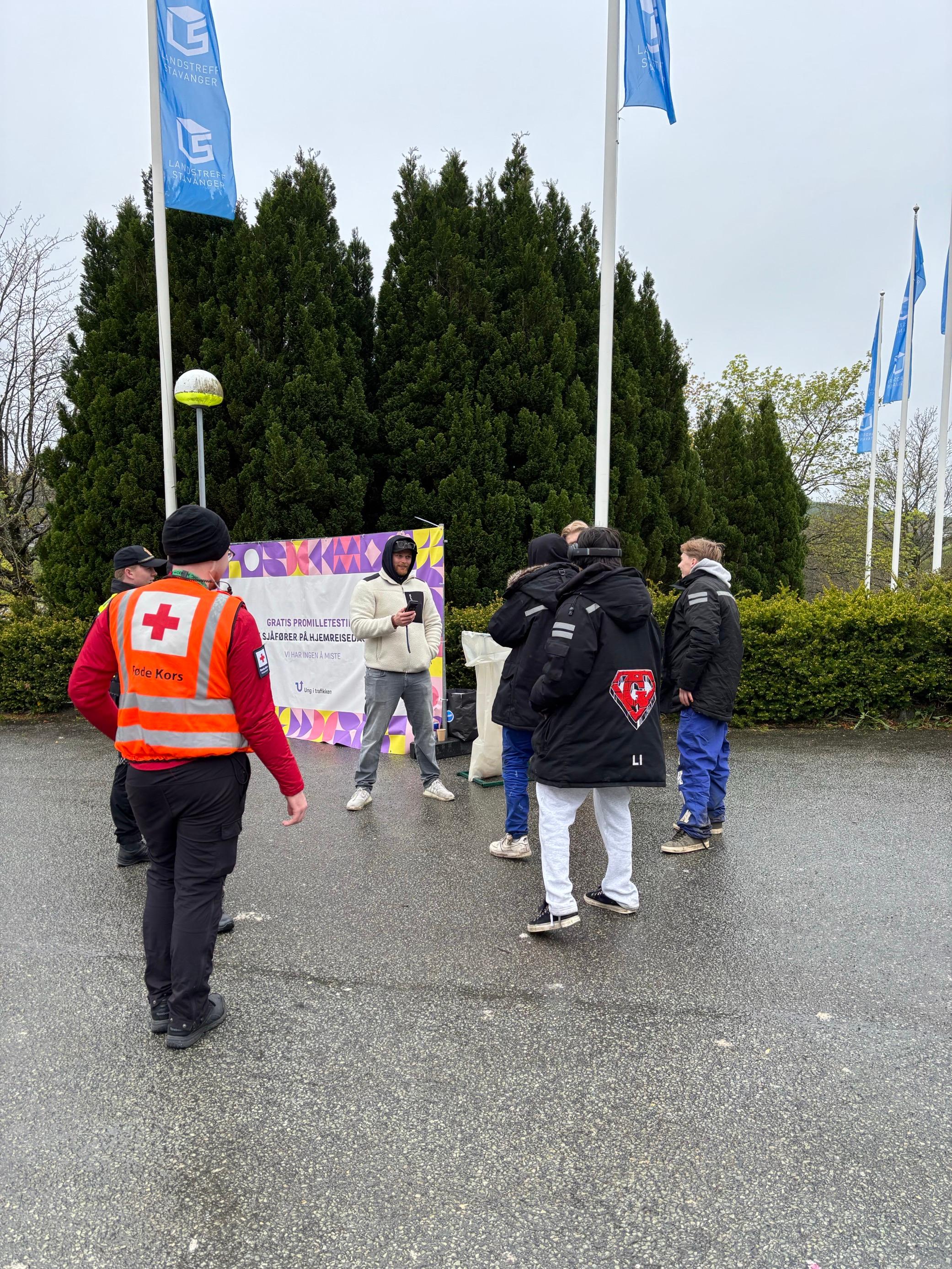 5 russ, Nikolay og en mann fra røde kors står utendørs foran et skilt der det står "Gratis promilletesting".