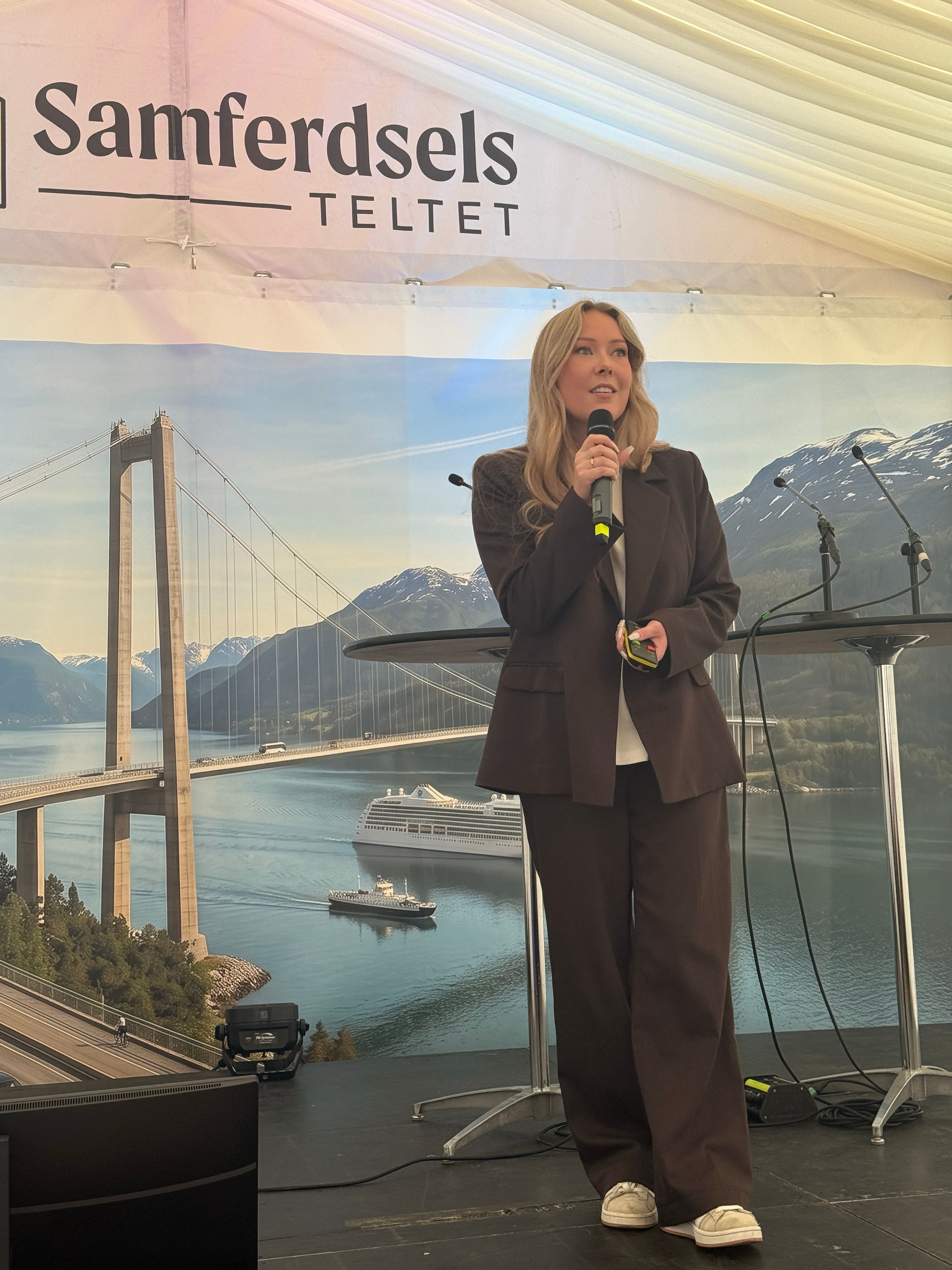 Ung dame står i bruk dress med mikrofon i hånden på en scene i samferdselsteltet på arendalsuka. Hun ser ut mot publikum.