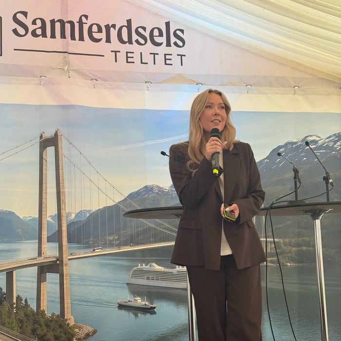 Ung dame står i bruk dress med mikrofon i hånden på en scene i samferdselsteltet på arendalsuka. Hun ser ut mot publikum.
