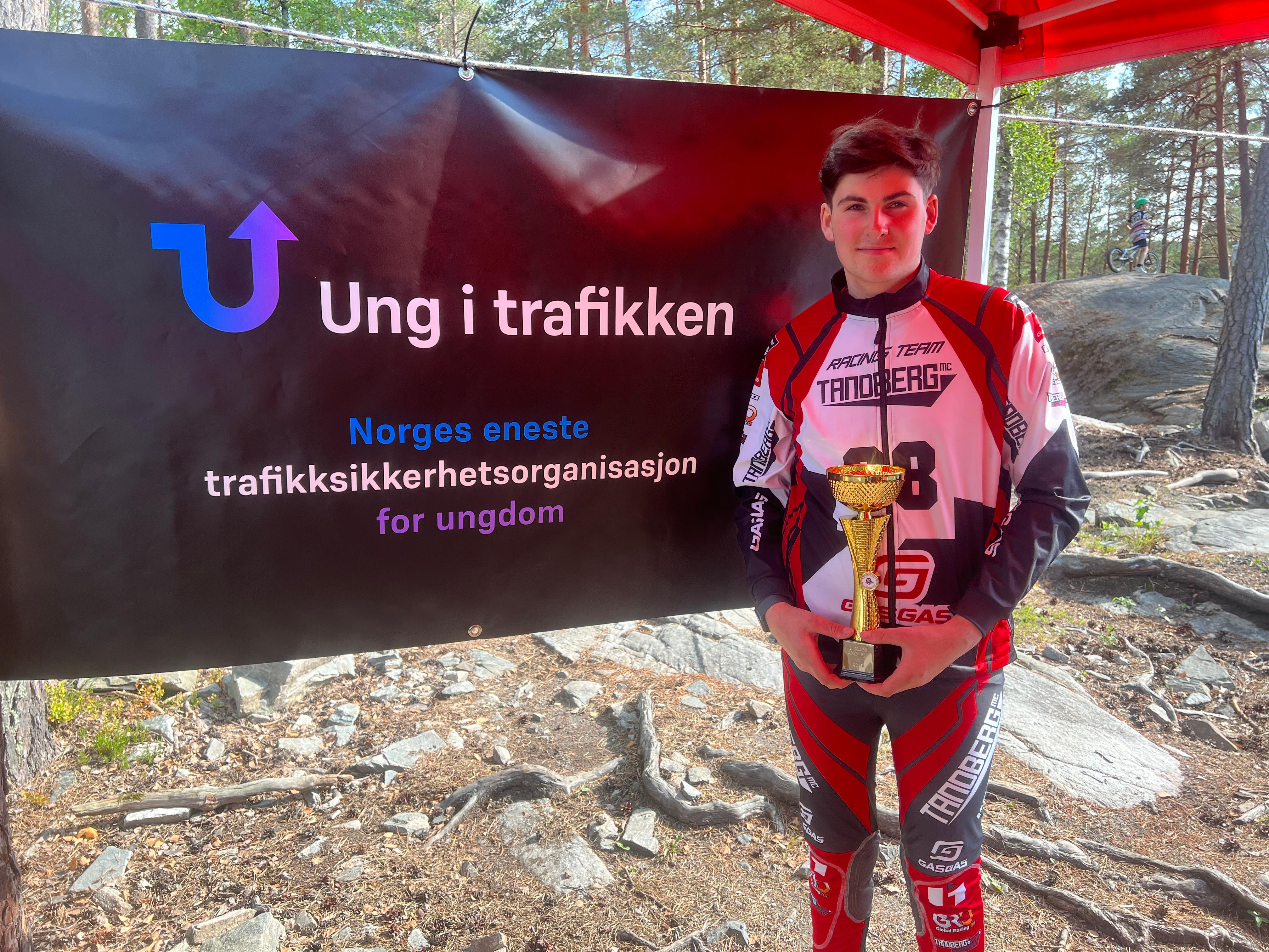 Bildet viser en ung trialfører i rød og hvit kjøredress med “Tandberg MC” og “GasGas” logoer. Han står under et rødt telt og holder en gullfarget pokal med begge hender, mens han smiler forsiktig mot kamera. Bak ham henger et stort svart banner med “Ung i trafikken” og teksten “Norges eneste trafikksikkerhetsorganisasjon for ungdom” i hvite og blå bokstaver. I bakgrunnen sees en skogkledd trialbane med en annen utøver som kjører motorsykkel over steiner og røtter.