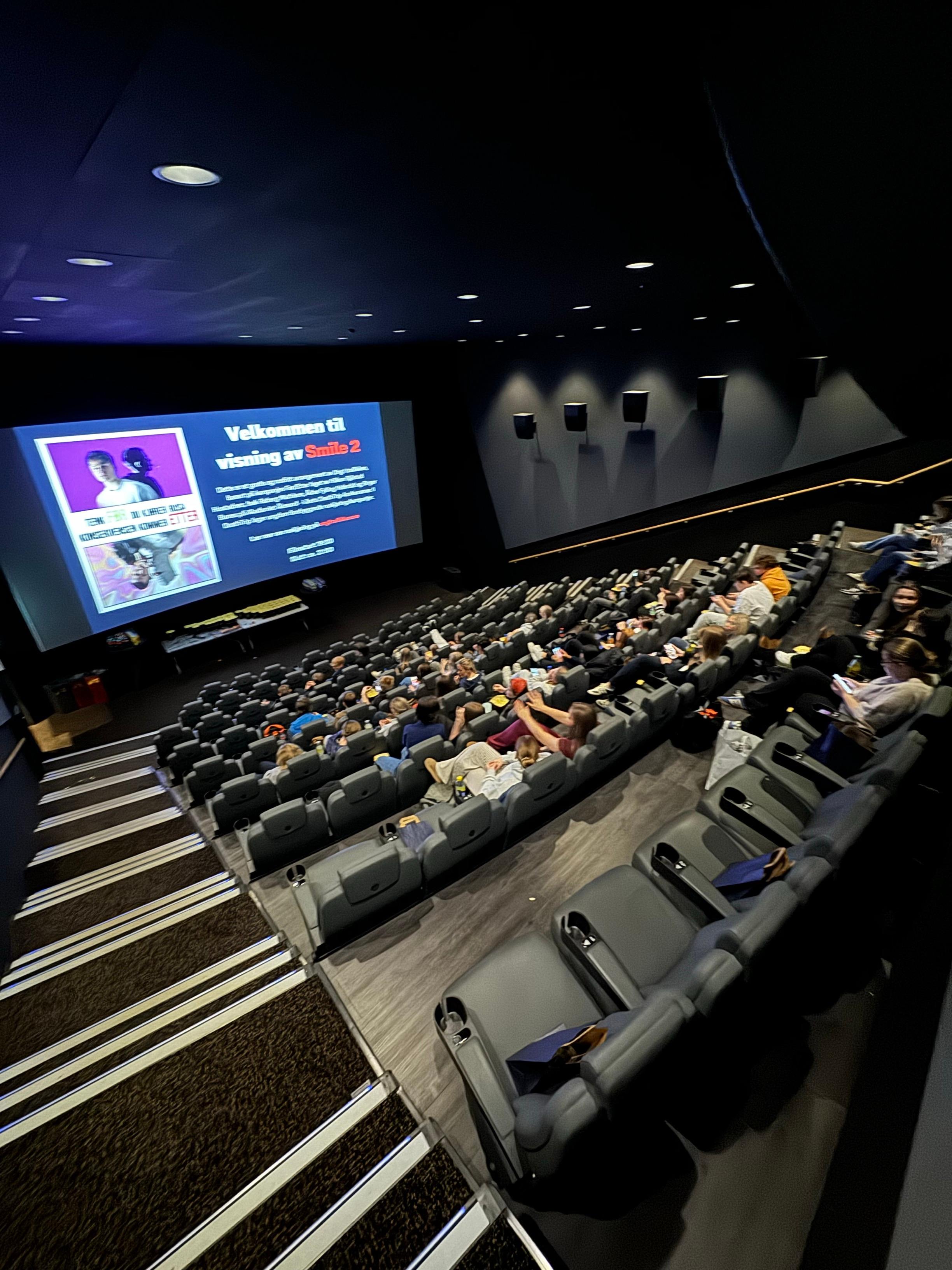 Bildet viser bilde fra kinosalen tatt fra øverste rad. Ungdommer som sitter i setene sine med popcorn, brus og goodiebags. På skjermen er en velkomstplakat som sier "Velkommen til kinovisning av Smile 2".