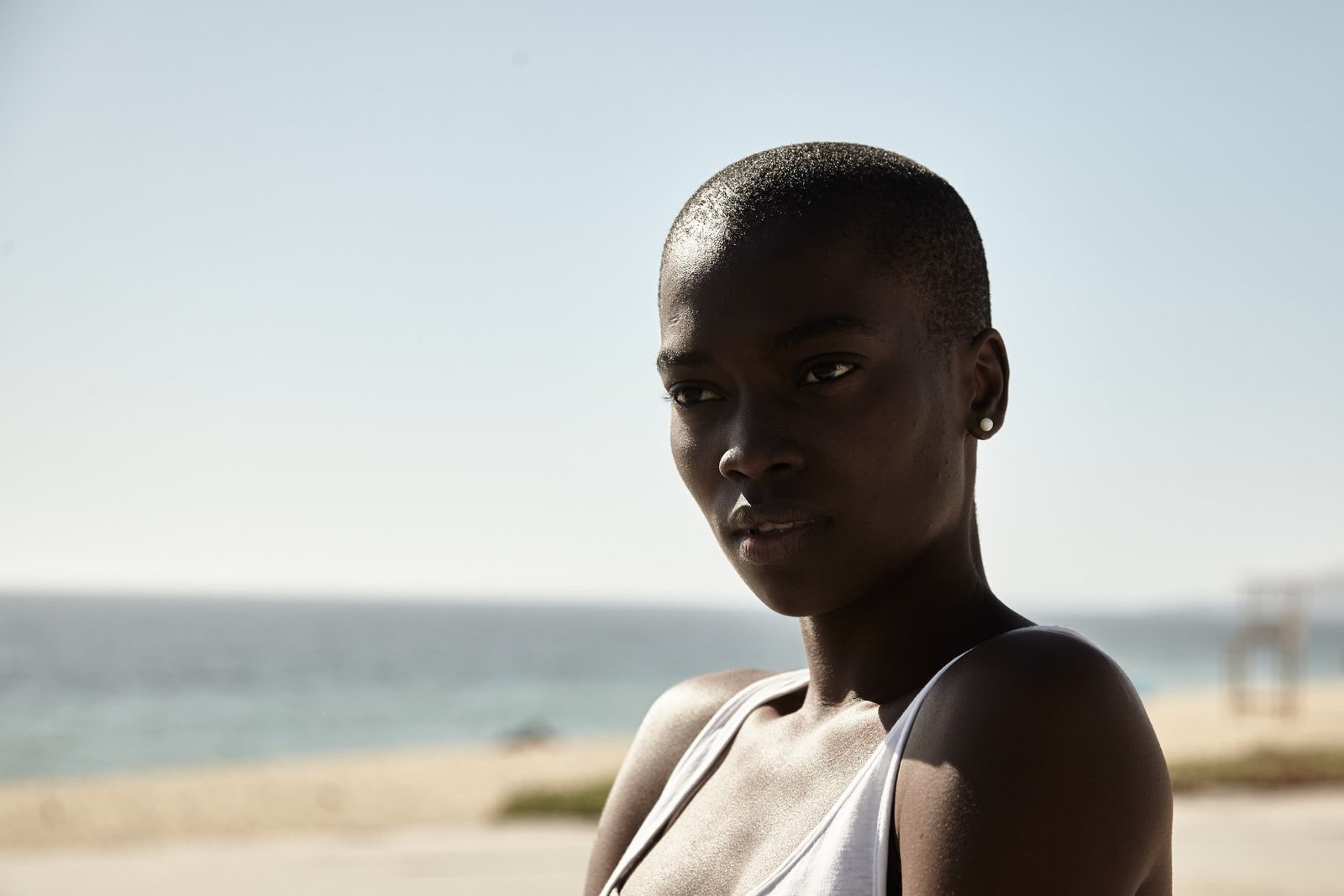 Black woman with closely shaved hair and a white tank top standing on a sandy beach, ocean and pale sky behind her