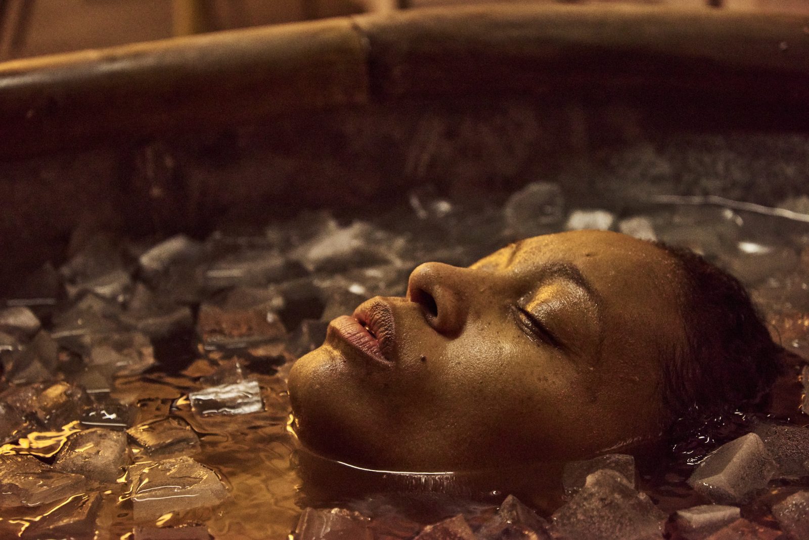Face floating eyes-closed in a wooden tub filled with ice, warm amber reflections in the dark water