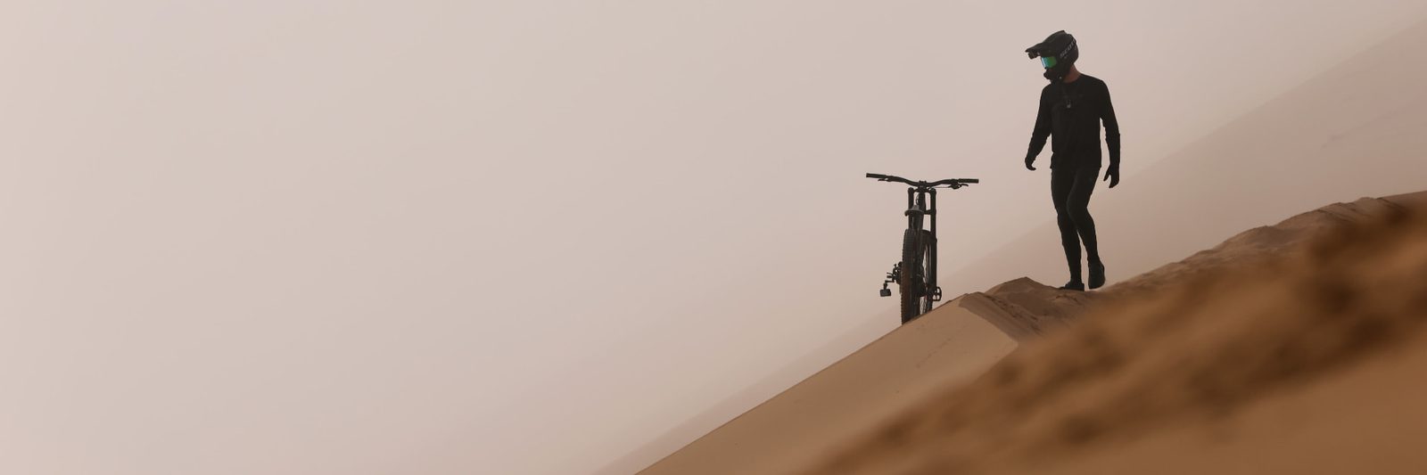 Cyclist in protective gear standing with a mountain bike on a misty, wind-swept dune ridge