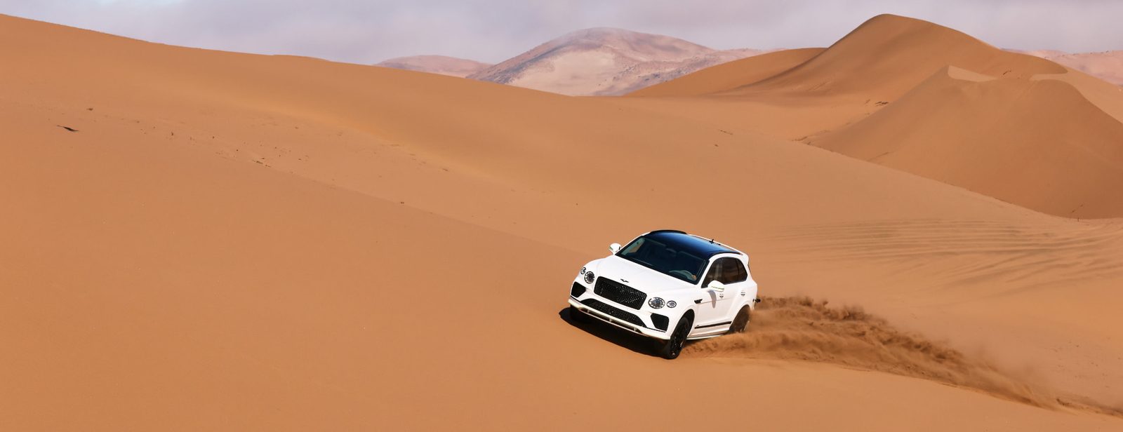 White Bentley Bentayga driving in a straight line across an expanse of desert sand dunes