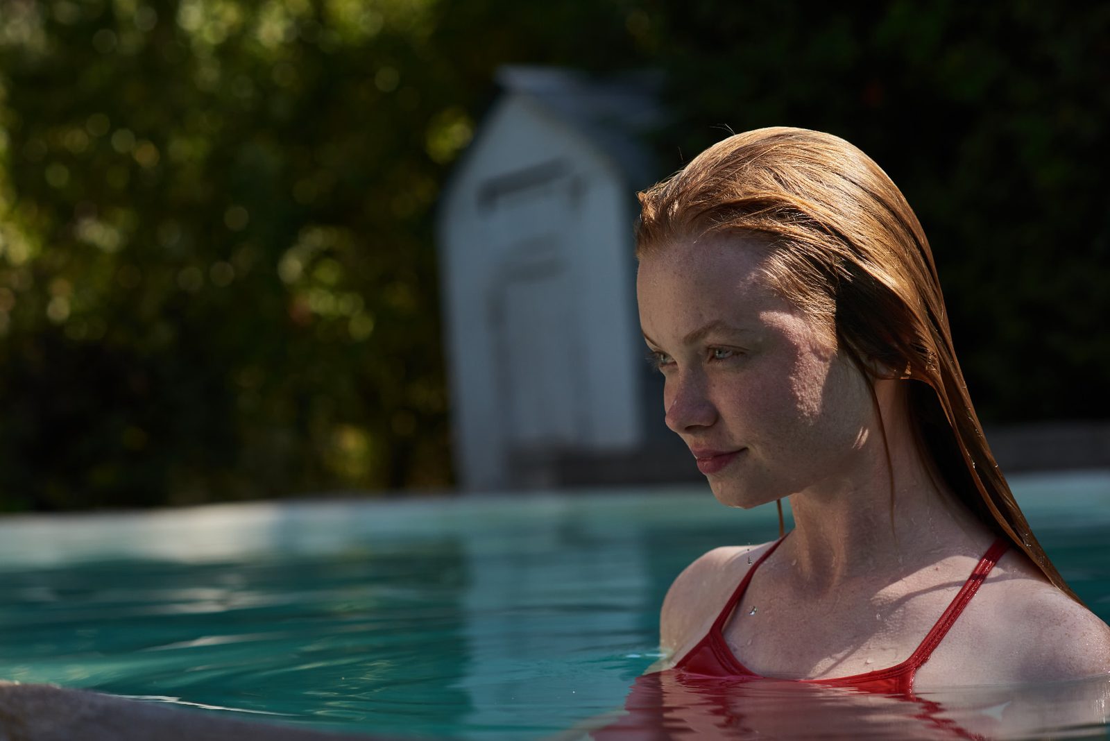 Red-haired woman submerged to her shoulders in a pool, framed against dark green foliage and bright sunlight