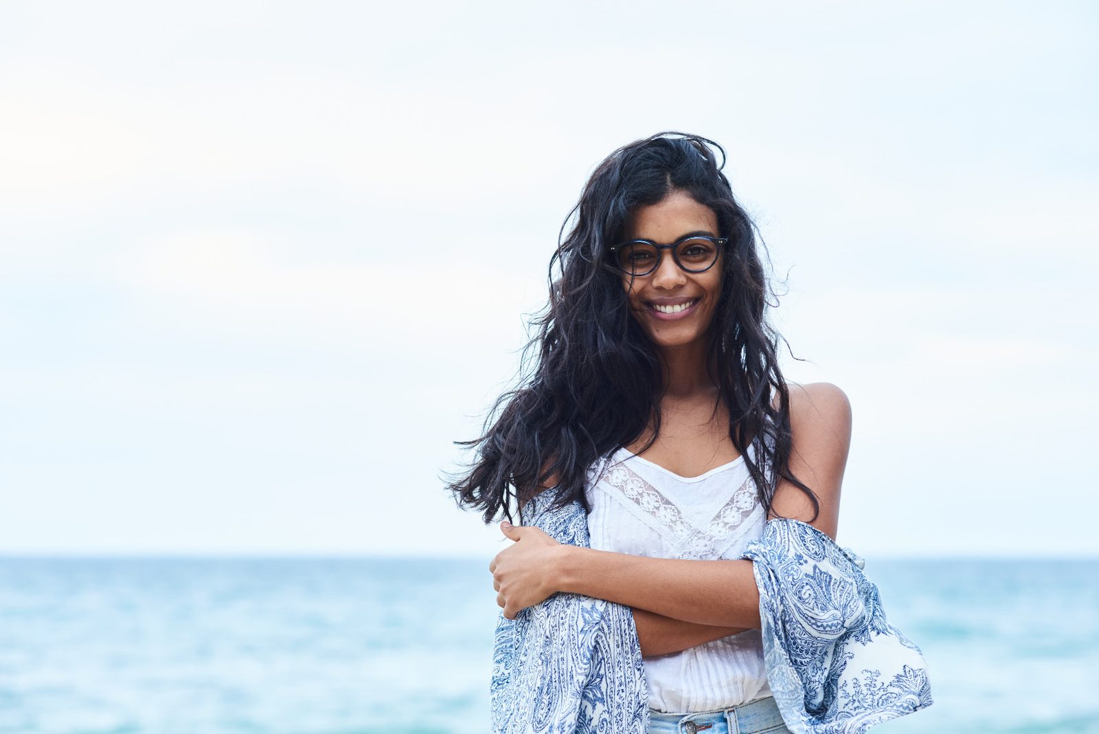 Smiling woman with long wavy hair and glasses on the coast, a blue-and-white patterned wrap draped over her arms