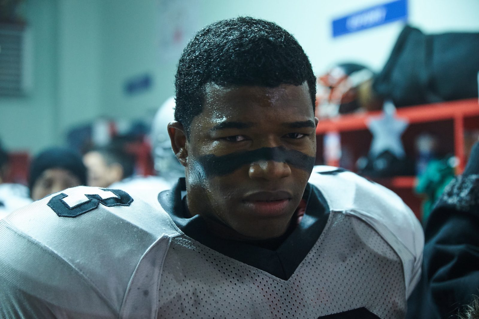 Football player in shoulder pads and eye grease staring hard in a locker room with red lockers behind him