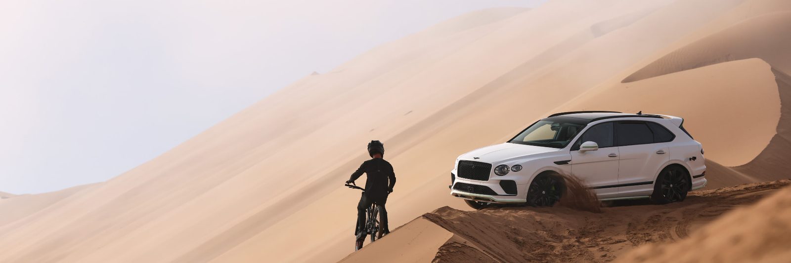 Mountain biker and white Bentley Bentayga cresting a line of enormous sand dunes, sand kicking up behind the vehicle
