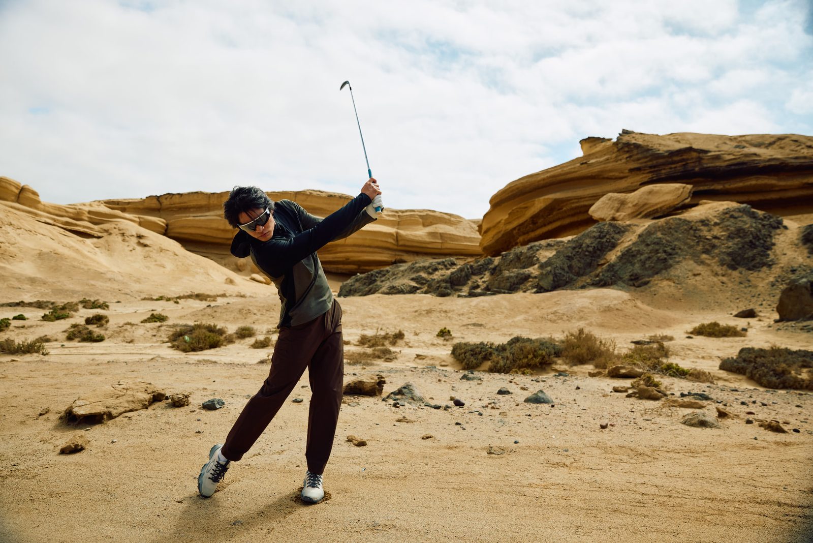 Golfer in shield sunglasses completing a swing follow-through on a desert plain ringed by sandstone ridges