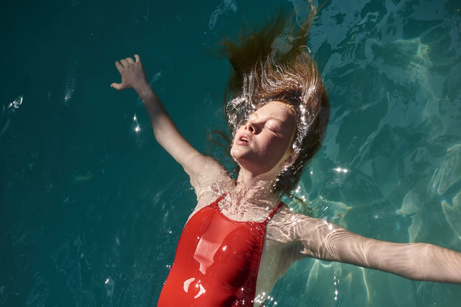 Young woman with copper hair floating on her back in a sunlit turquoise pool, arms outstretched, wearing a red swimsuit