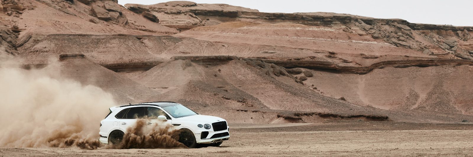 White Bentley Bentayga sliding sideways across an arid plain, spraying dirt toward the camera