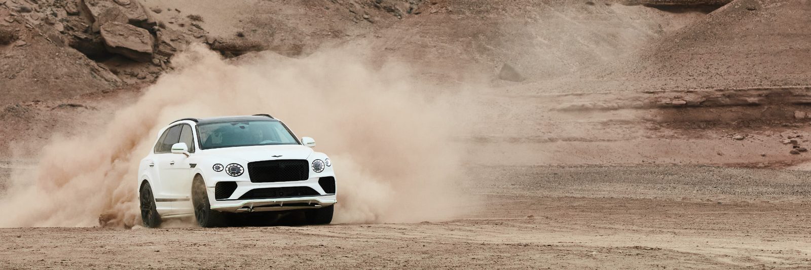 White Bentley Bentayga drifting across a desert canyon, throwing a towering cloud of dust behind it