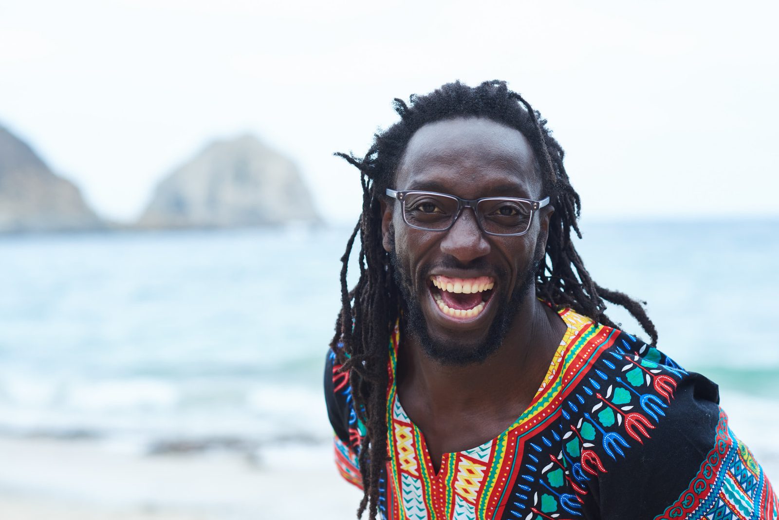 Man with dreadlocks and glasses laughing openly on a beach, wearing a black dashiki with red, yellow and turquoise geometric patterns