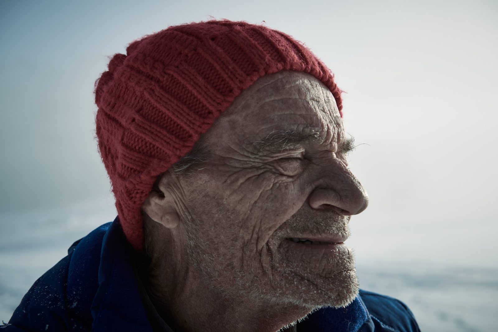 Close-up of the same elderly man in a red beanie, eyes closed in a warm smile against pale snow