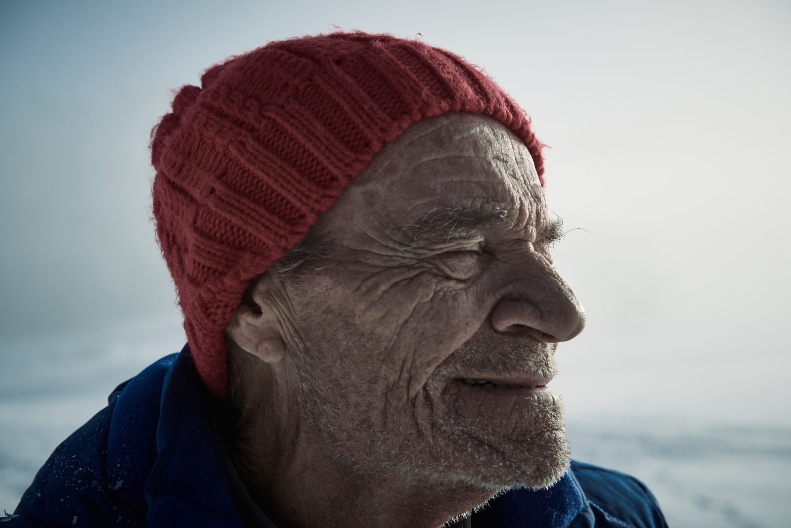 Close-up of the same elderly man in a red beanie, eyes closed in a warm smile against pale snow
