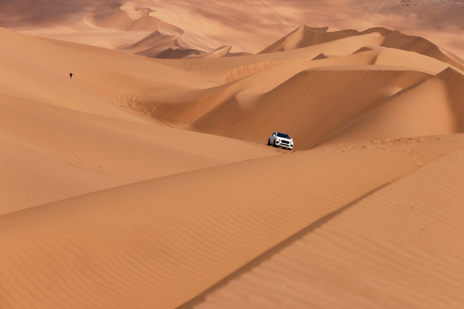 White Bentley Bentayga crossing vast desert dunes, a drone flying above it to the upper left