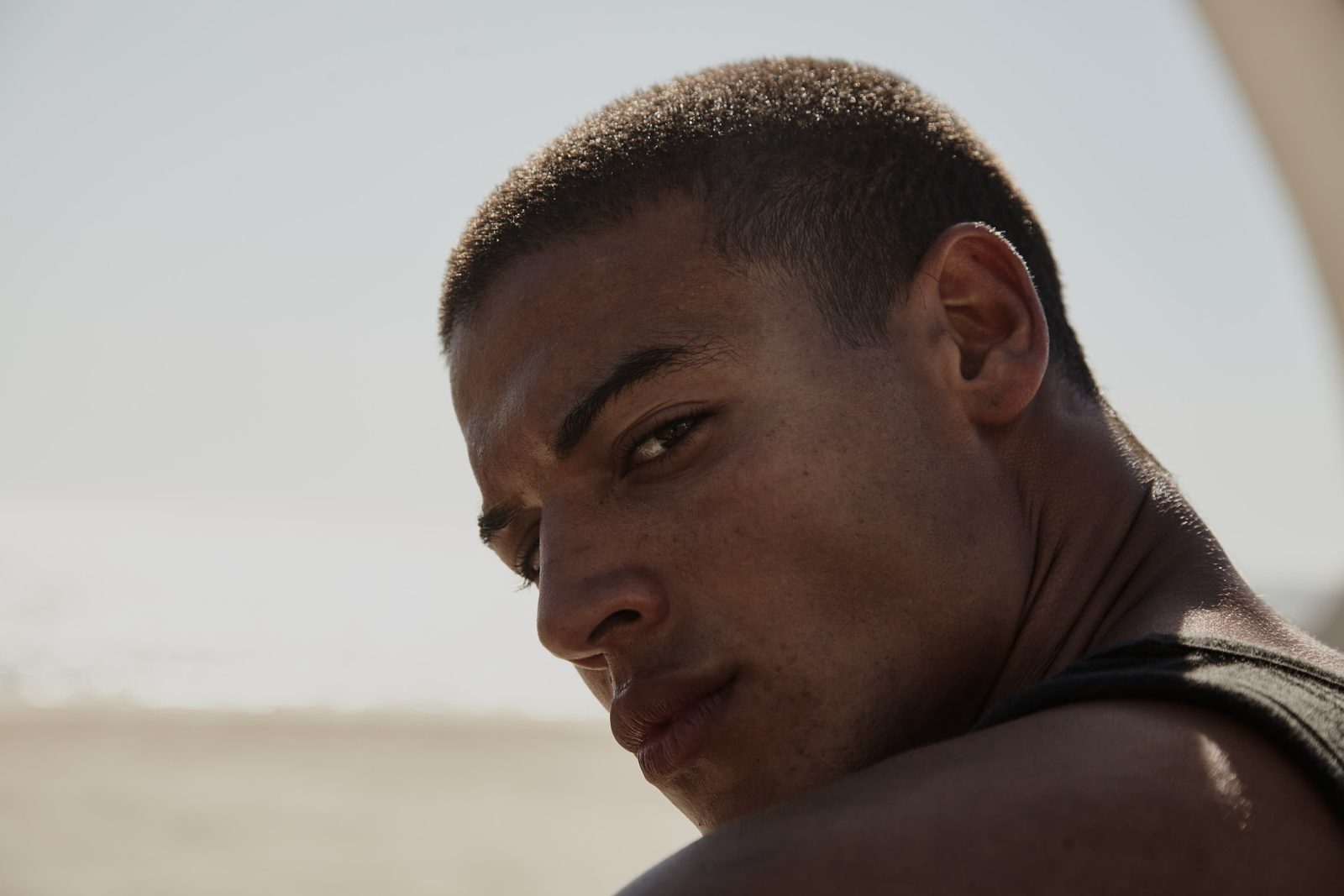 Extreme close-up of a young man with a buzz cut looking back over his shoulder toward the camera, bright beach behind him