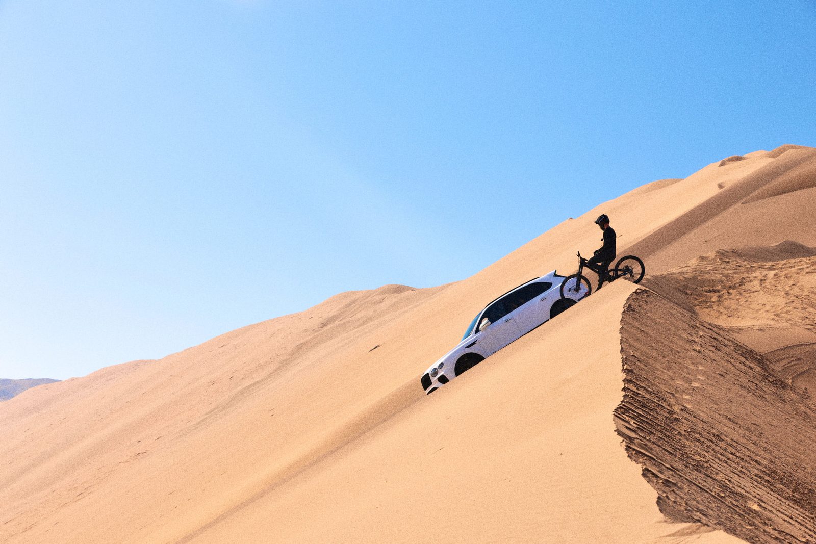 White Bentley Bentayga and a mountain biker stopped together on the crest of a sand dune under a clear sky