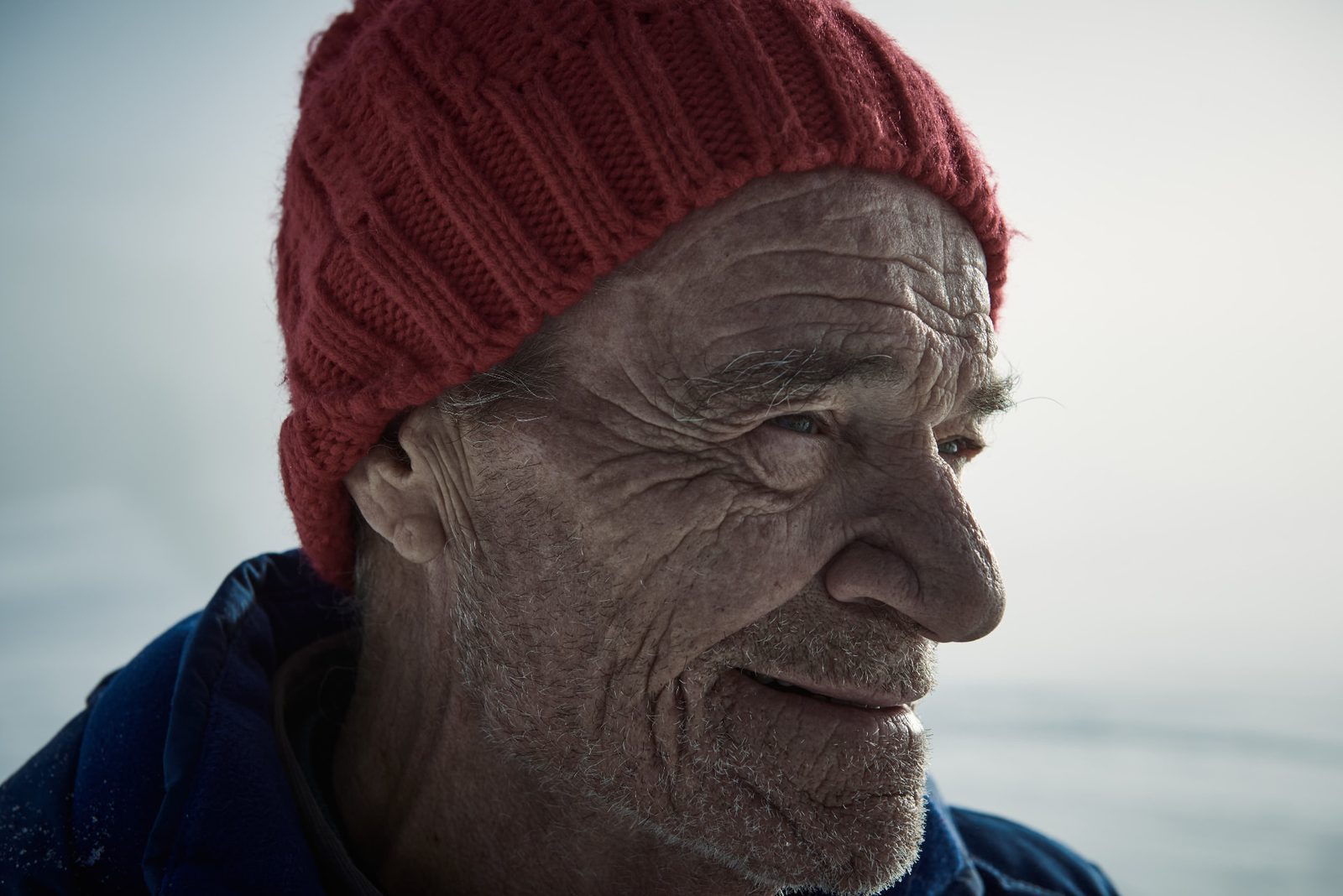 Close-up of an elderly weathered face in a red knit beanie, looking off to the side against a snow-covered landscape