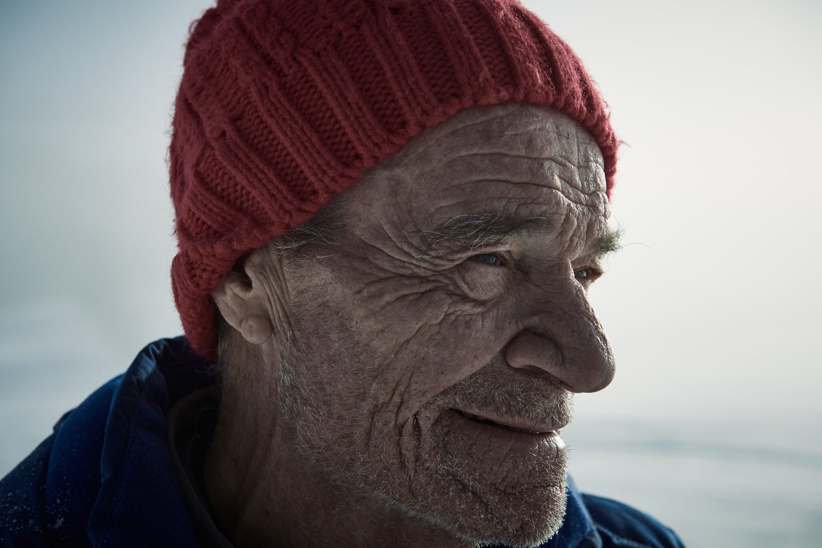 Close-up of an elderly weathered face in a red knit beanie, looking off to the side against a snow-covered landscape