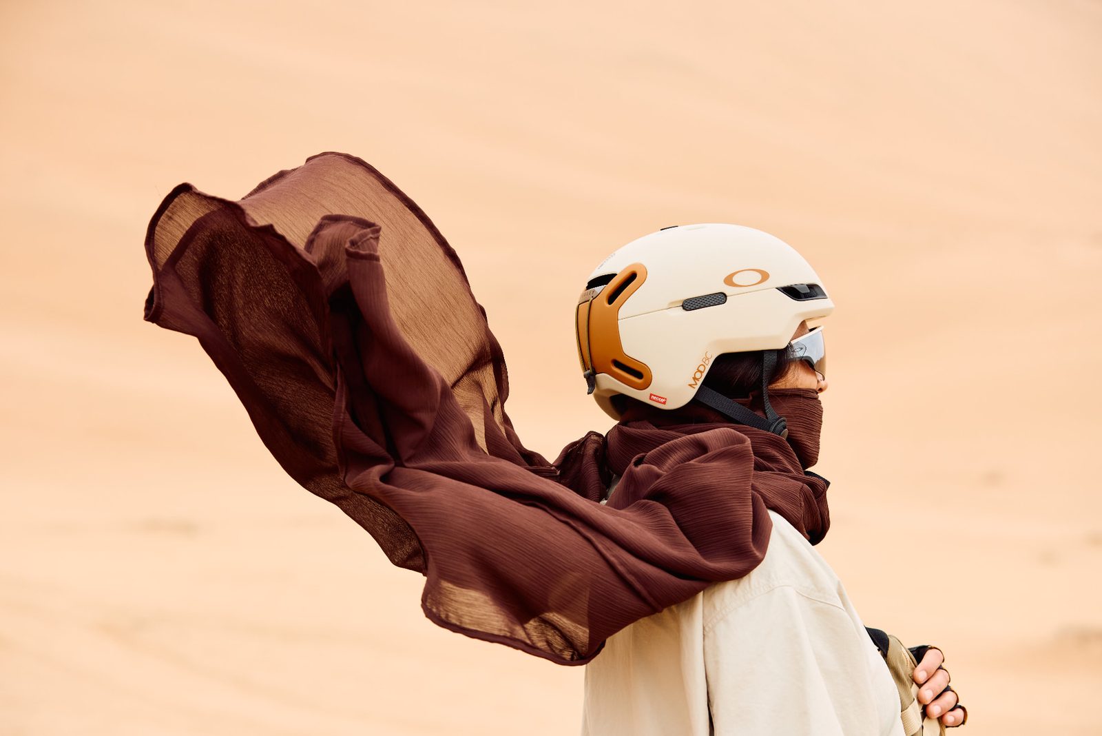 Sandboarder in profile on a dune, helmet and sunglasses, long brown scarf streaming in the wind