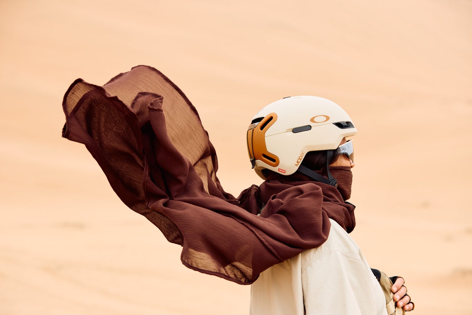 Sandboarder in profile on a dune, helmet and sunglasses, long brown scarf streaming in the wind