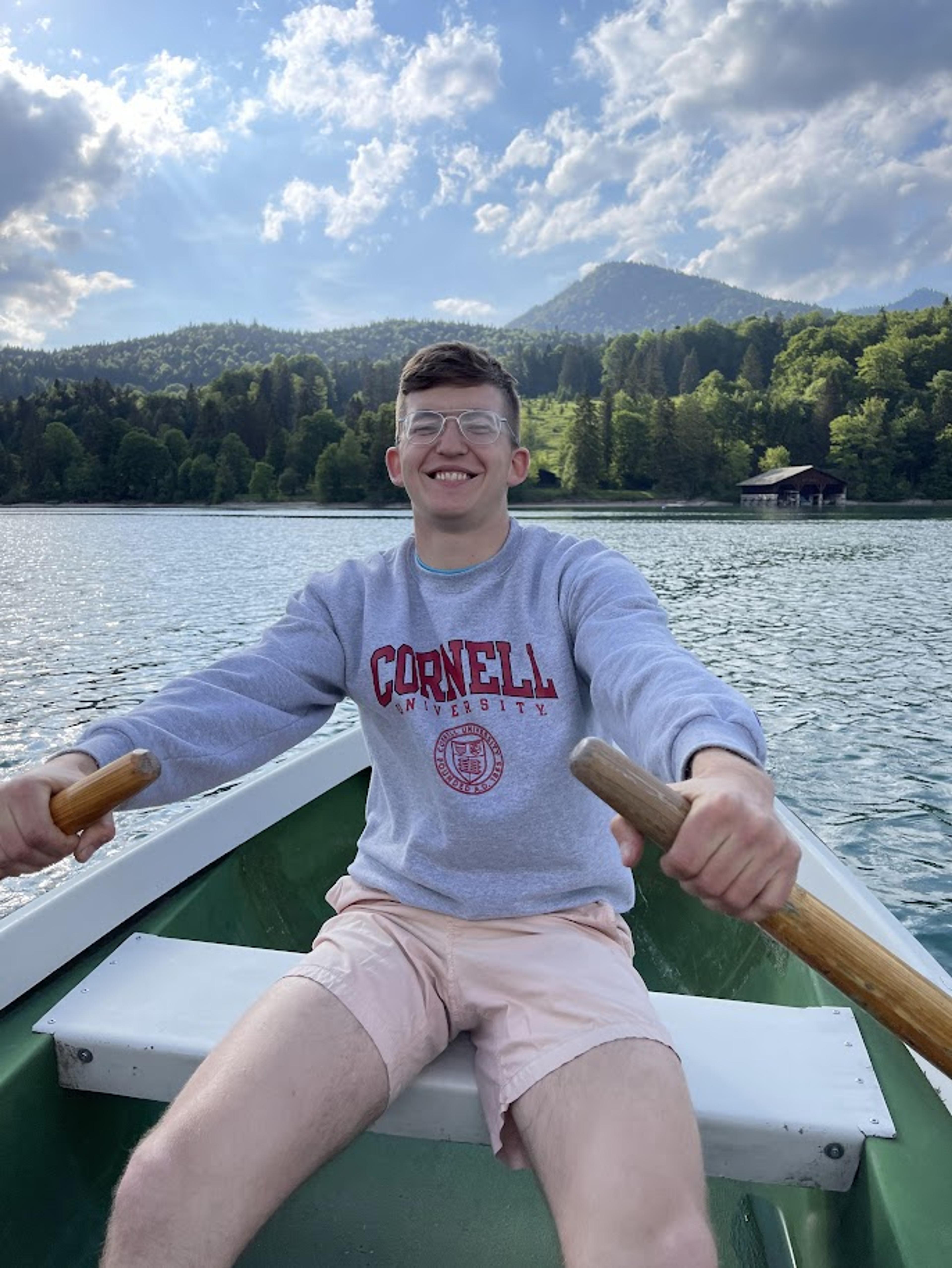 A young man smiling at the camera, rowing a boat on a lake