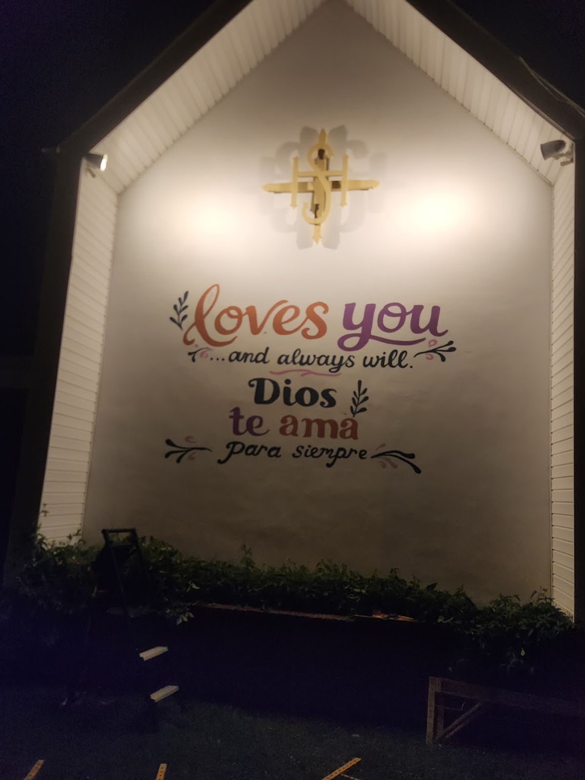 The God Loves You mural on the white gable end of a church, floodlit at night beneath a gold cross. Hand-lettered script reads "loves you …and always will." above the Spanish "Dios te ama para siempre."
