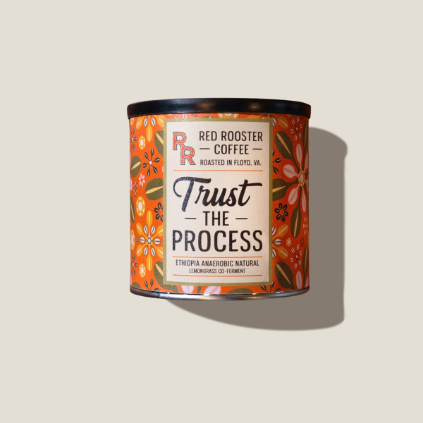 A patterned orange tin of Red Rooster Coffee labeled “Trust the Process” sits against a plain beige background, casting a soft shadow; the image feels warm and understated.
