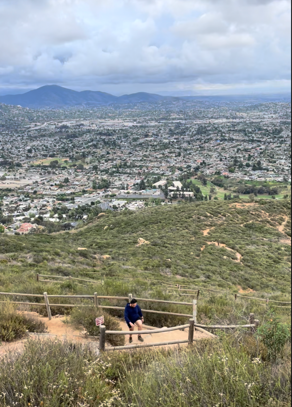 Hiker climbing the wood-railed switchback trail near the summit of Cowles Mountain, with sweeping views over San Diego's neighborhoods and distant mountains
