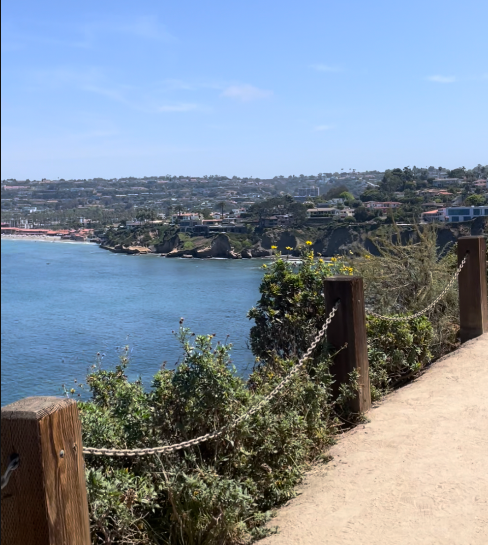 Sandy cliffside path with a wood-post-and-chain railing along the Coast Walk Trail in La Jolla, overlooking a turquoise cove and the coastline