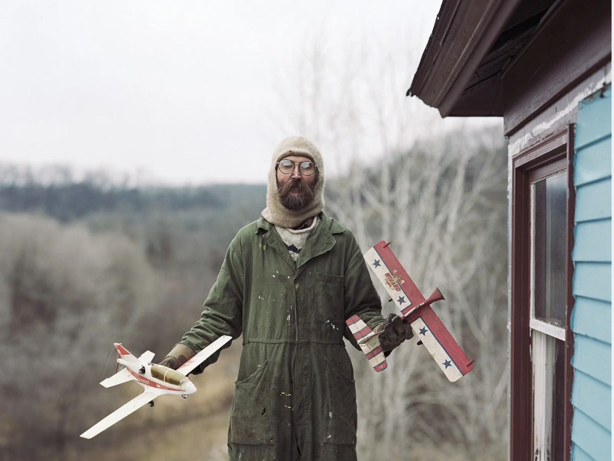 Alec Soth holding two model airplanes outdoors beside a wooden building