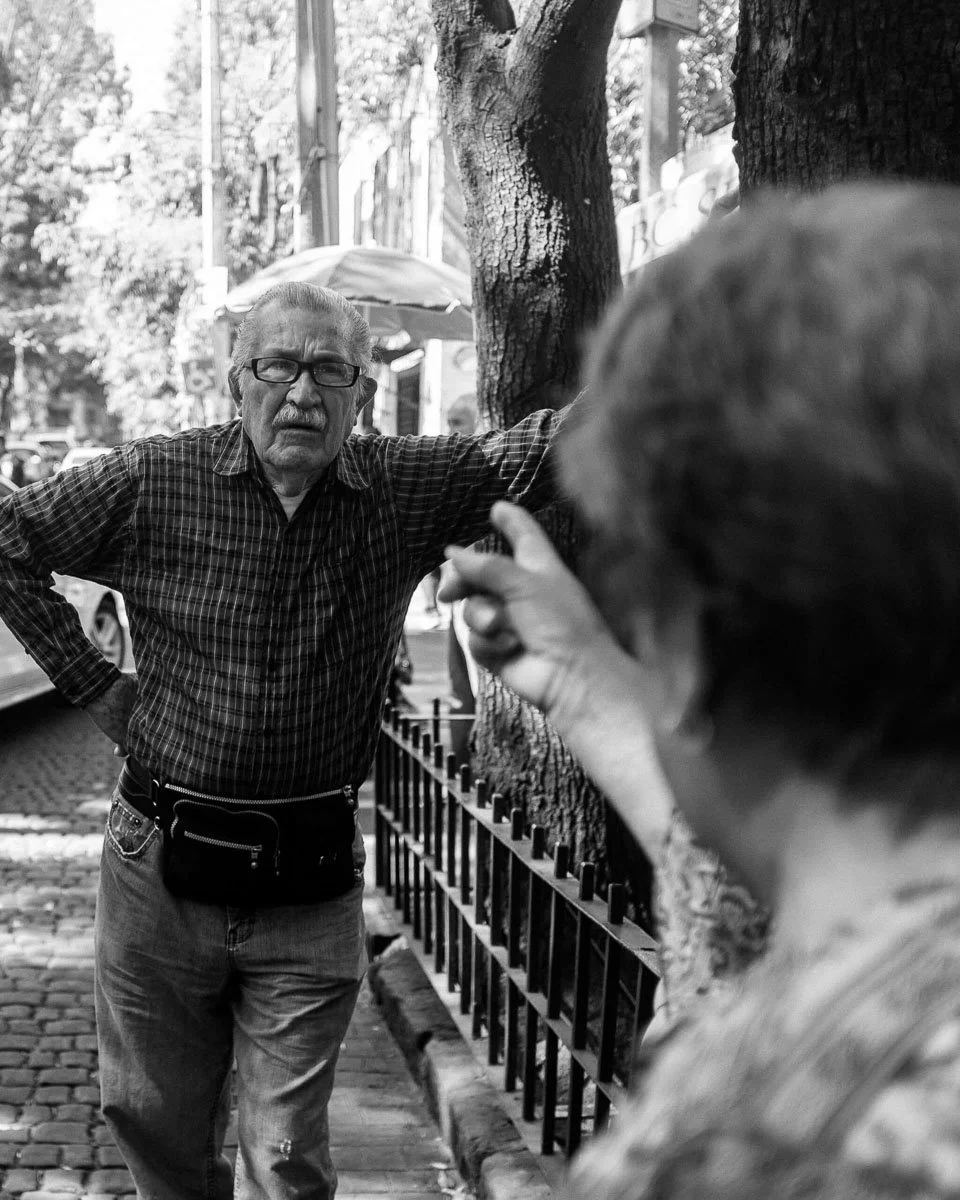 Black-and-white Mexico City street photograph of an older man gesturing beside a fence