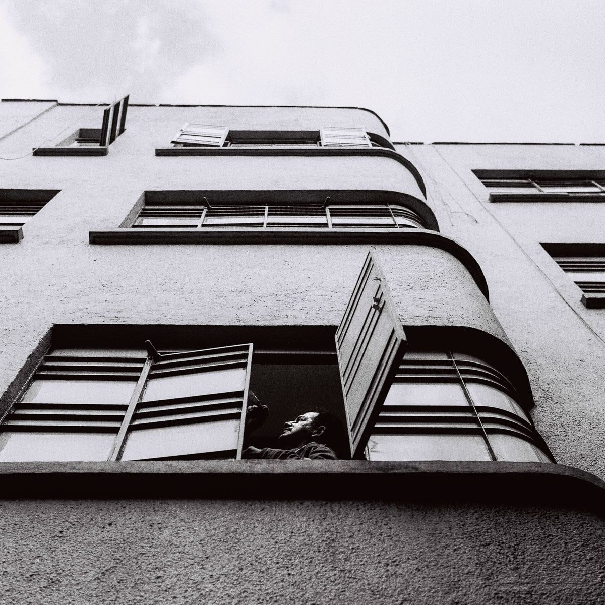 Black-and-white analog street photograph of a man looking from an apartment window.