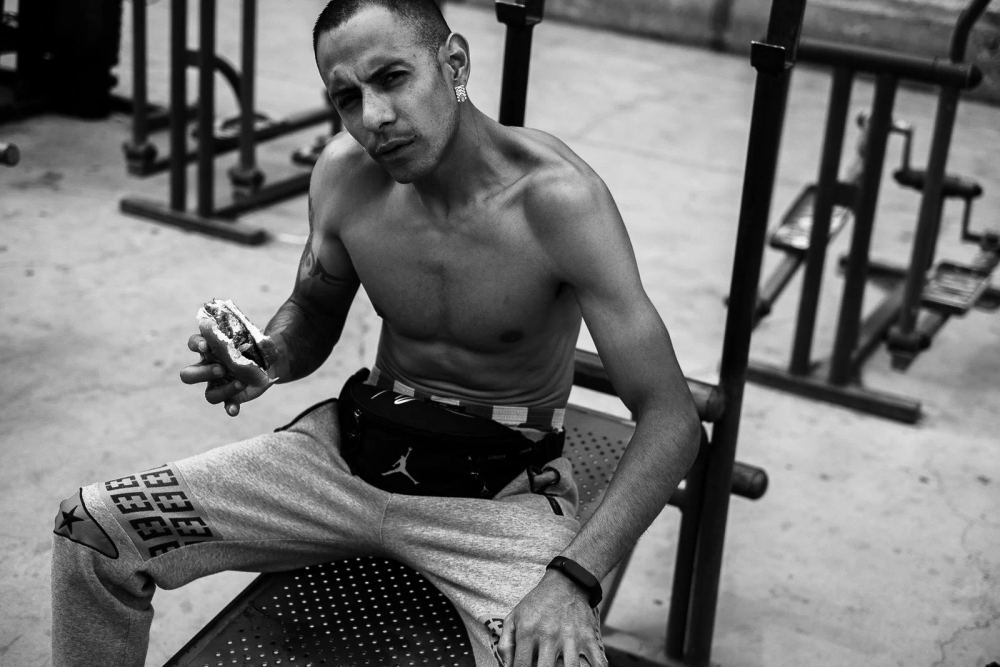 Black-and-white photograph of a man holding food while seated on outdoor exercise equipment