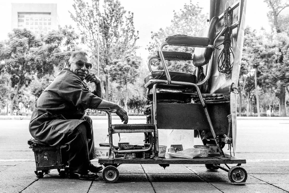 Black-and-white street portrait of a man seated beside an outdoor shoeshine chair