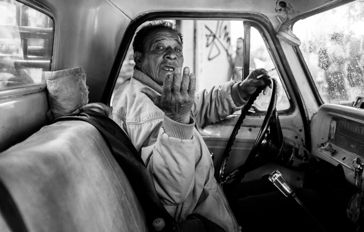 Black-and-white portrait of a truck driver gesturing from behind the steering wheel