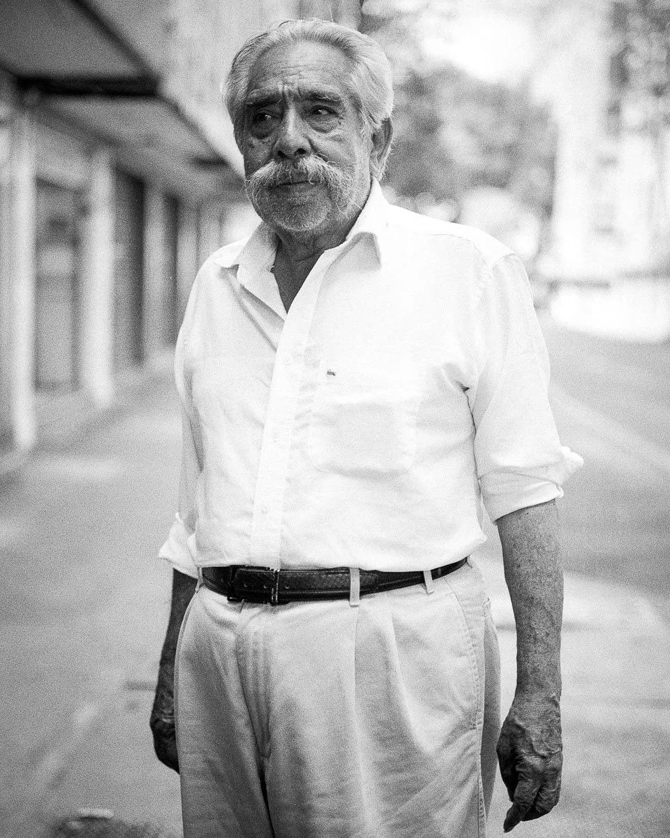 Black-and-white street portrait of an older man in a white shirt and light trousers