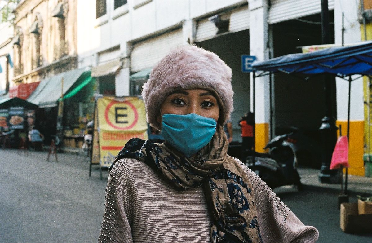 Color film street portrait of a woman in a pink hat and blue face mask.
