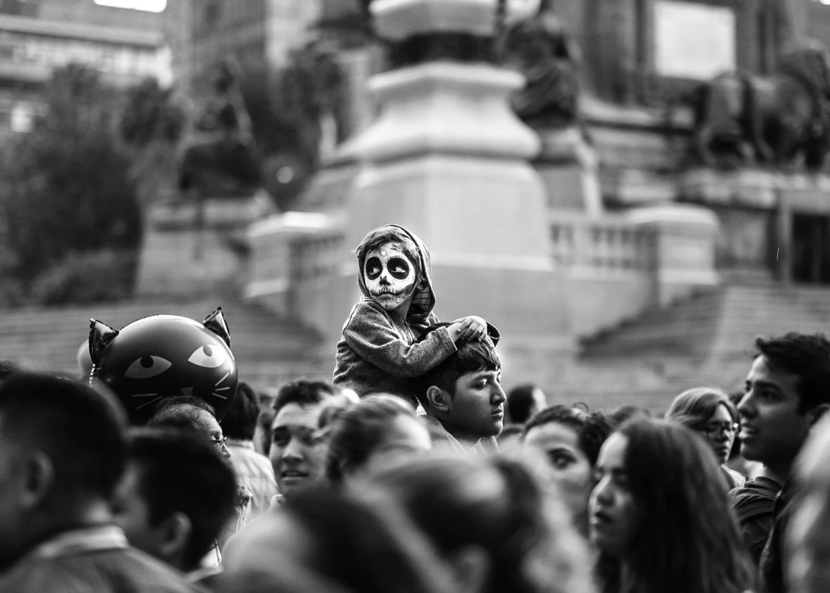 A child with skull face paint sitting above the crowd during Day of the Dead in Mexico City