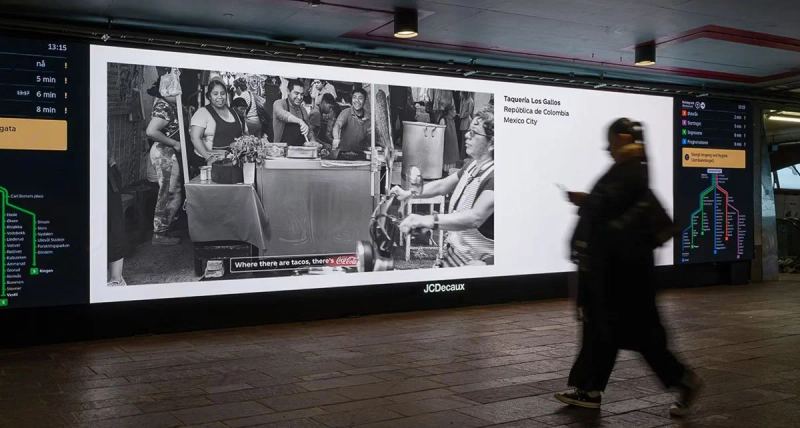 A commuter passes a wide illuminated display of Coca-Cola campaign photographs