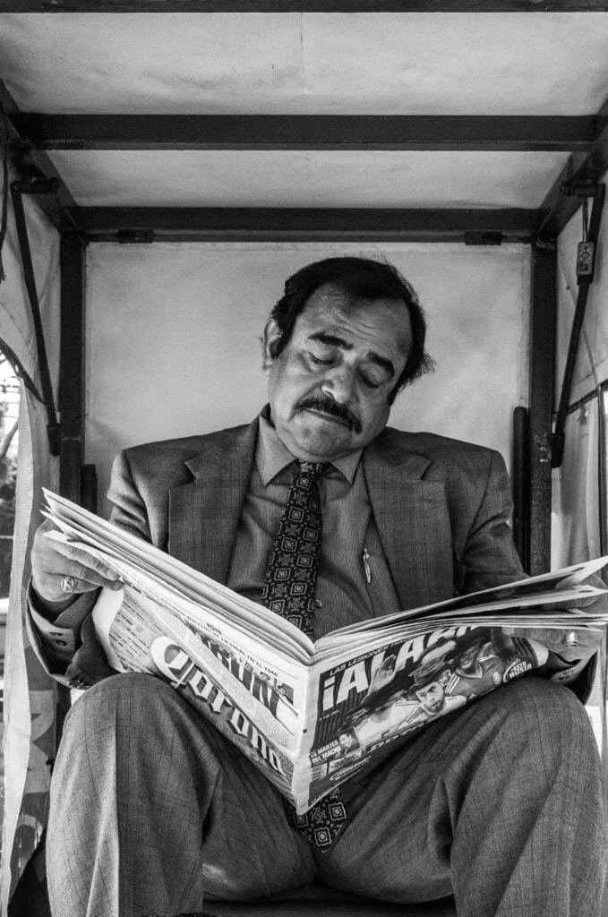 Black-and-white portrait of a man reading a newspaper in a narrow street booth