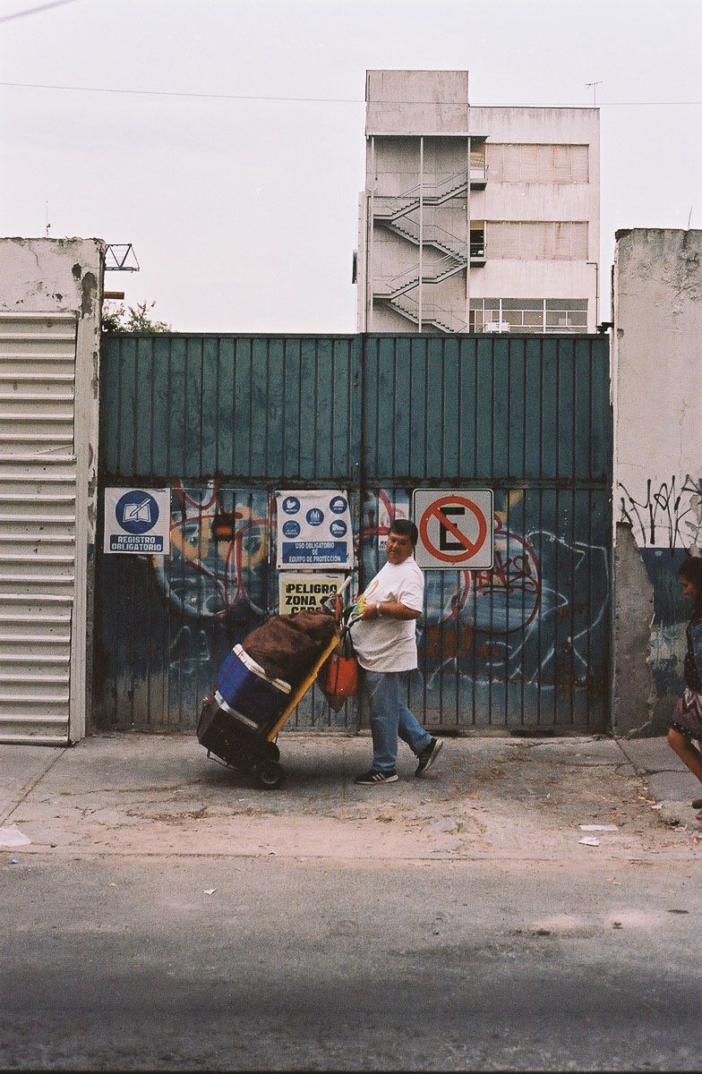 Color analog street photograph of a man pushing a loaded trolley past an industrial gate.
