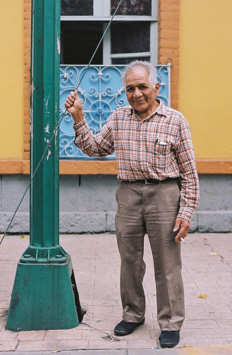 Color film street portrait of an older man holding a wire beside a green lamppost.